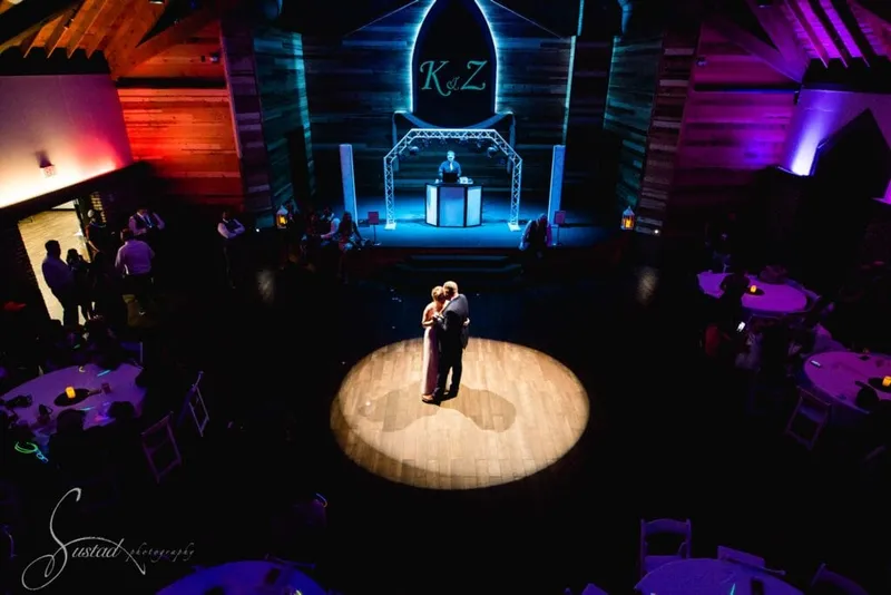 Couple sharing a first dance under a spotlight in a wood-beamed reception hall with colorful uplighting and a DJ booth