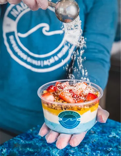 Hand holding an açaí bowl topped with strawberries, granola, and shredded coconut being sprinkled from a spoon