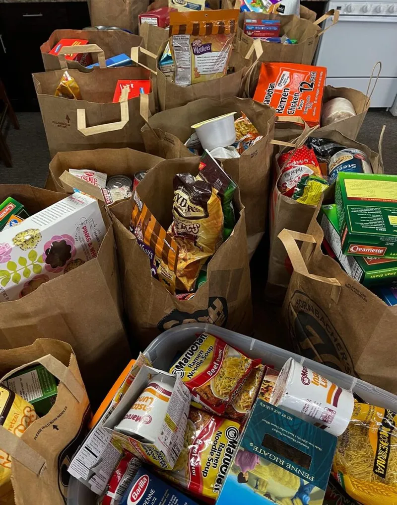 Brown paper grocery bags overflowing with ramen, pasta, snacks, and canned goods for a food drive