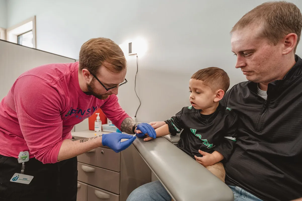 Gloved phlebotomist draws blood from a young boy's arm while an adult holds him in a clinic exam room