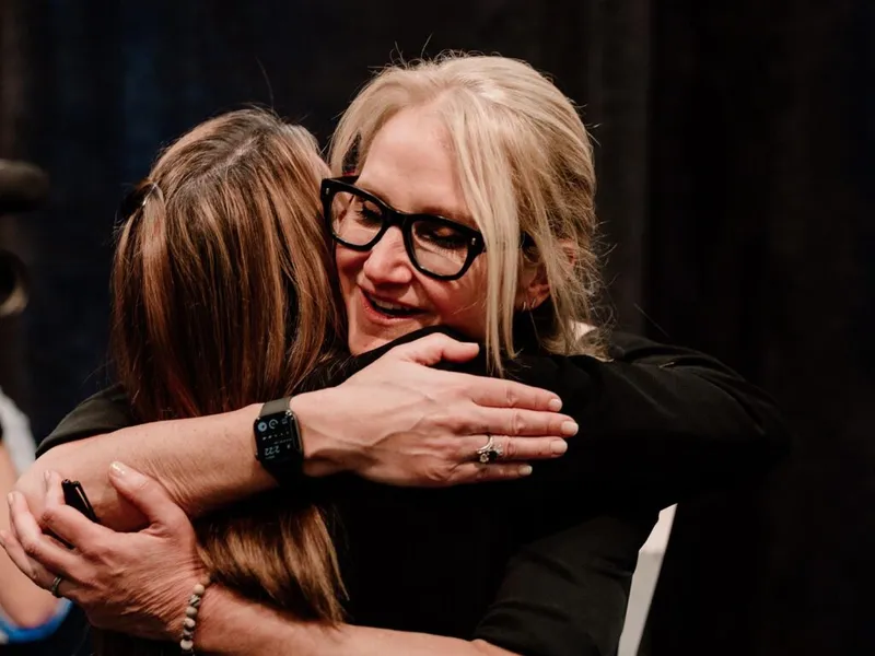 Two women in black tops embracing warmly at an event, one wearing black-framed glasses and a smartwatch