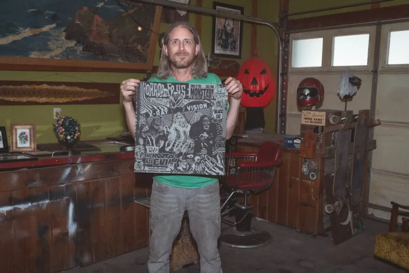 Long-haired man in green shirt holding a large horror-themed illustrated poster in a eclectic garage studio