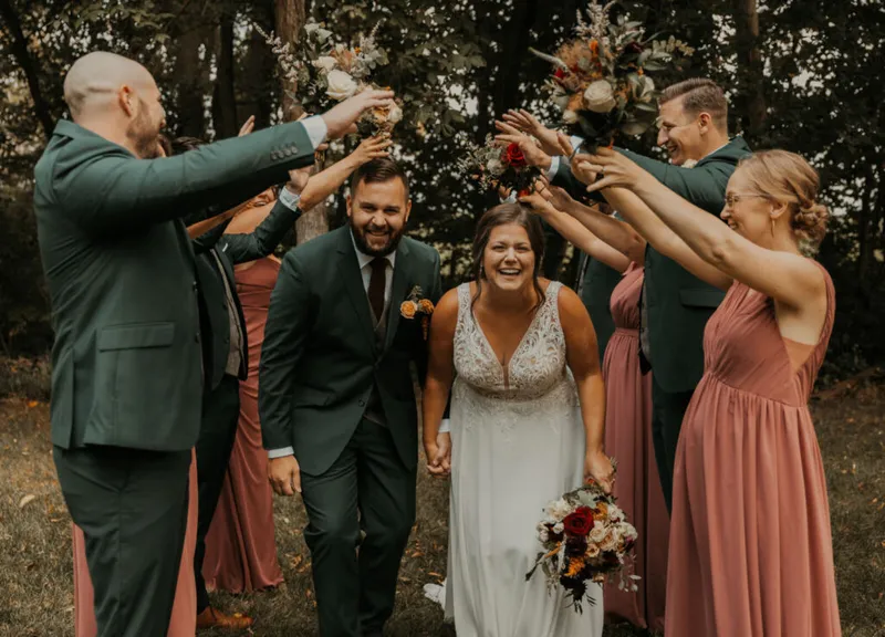 Laughing bride and groom walk hand-in-hand under an arch of bouquets raised by their wedding party