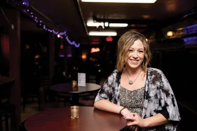 Smiling woman with blonde hair seated at a round table in a dimly lit bar or comedy club