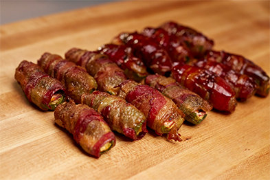 Bacon-wrapped jalapeño poppers on a wooden cutting board, some glazed with red sauce