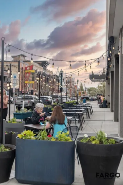 Two women dining on a downtown sidewalk patio under string lights at dusk