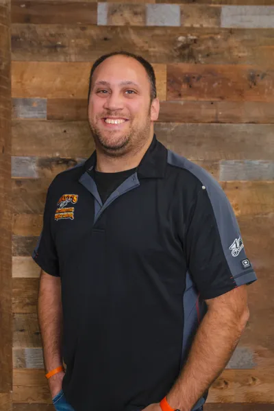 Smiling man in a black and gray logo polo shirt standing before a reclaimed wood wall