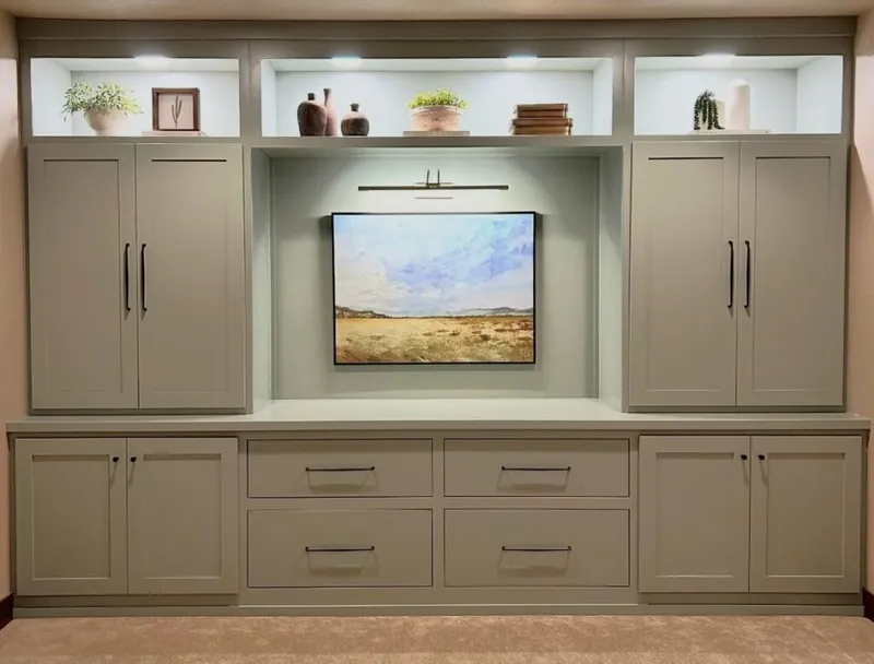 Built-in sage green cabinetry with lit shelves, drawers, and a landscape painting in the center niche
