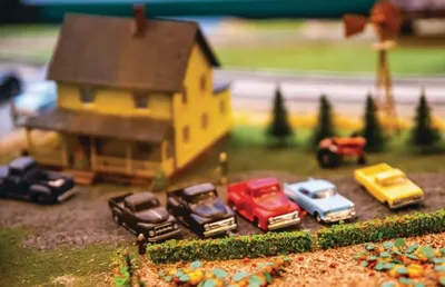 Miniature model cars parked near a small yellow farmhouse with trees
