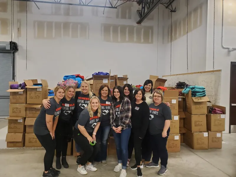 Group of volunteers in SCHEELS Helping Hands shirts posed in front of stacked donation boxes in a warehouse