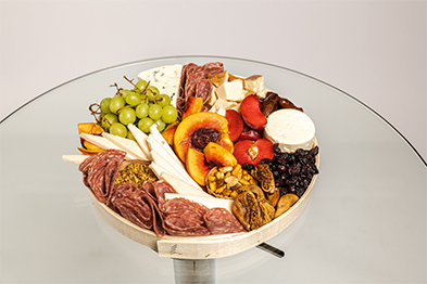 Round wooden charcuterie board with salami, cheeses, grapes, sliced peaches, strawberries, and dried fruits