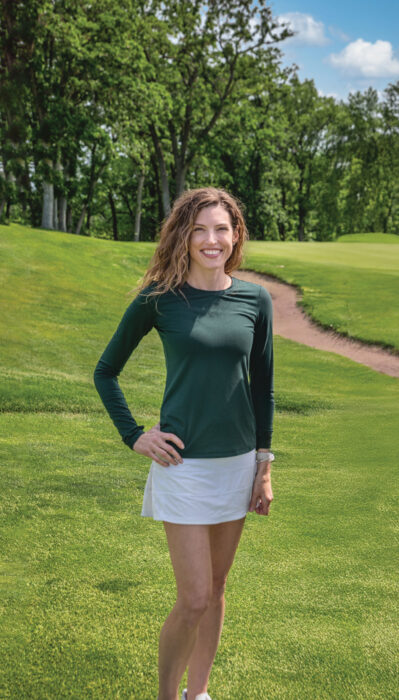 Smiling woman in dark green long-sleeve top and white skirt standing on a golf course fairway