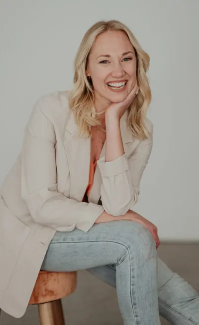 Smiling blonde woman in a cream blazer and jeans seated on a stool, chin resting on hand