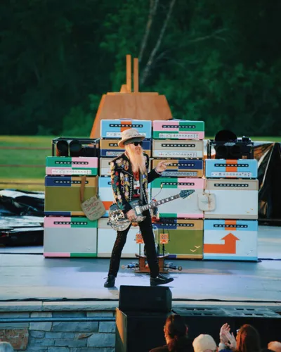 Guitarist with long beard and hat performs on outdoor stage backed by colorful stacked amplifiers