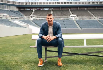 Smiling man in blue suit and tan boots seated on a bench at an empty stadium