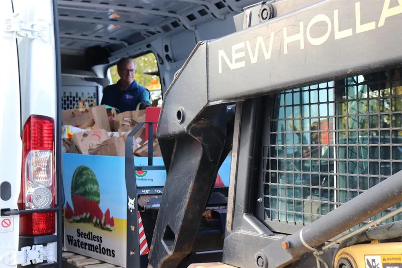 Man loading brown paper bags into a van beside a New Holland skid steer loader
