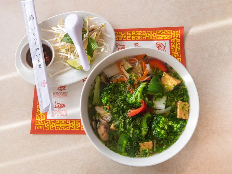 Overhead view of a vegetable pho bowl with tofu, broccoli, and herbs beside a bean sprout garnish plate