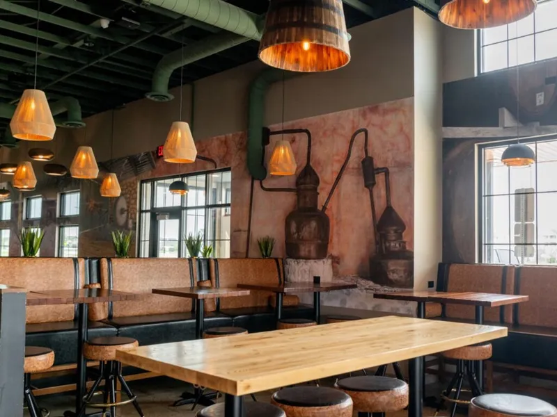 Restaurant dining room with wood tables, cork stools, pendant lights, and a copper still mural on the wall