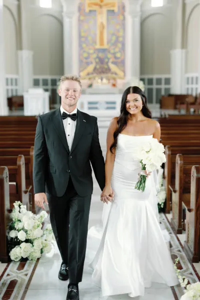 Smiling bride in strapless white gown and groom in black tuxedo walk hand-in-hand down a church aisle