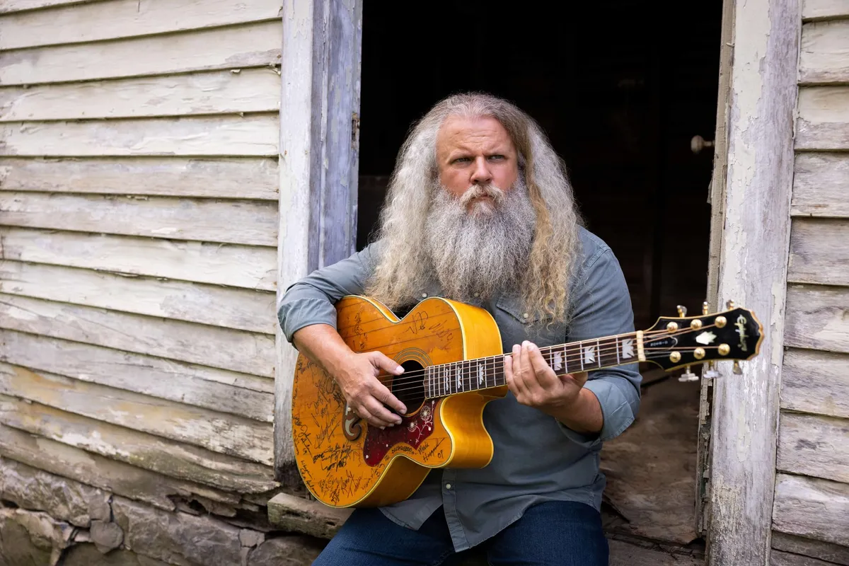 Long-haired bearded man playing a signature-covered acoustic guitar in a weathered barn doorway