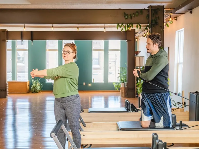 Two people on Pilates reformers in a bright studio with wood floors and hanging plants