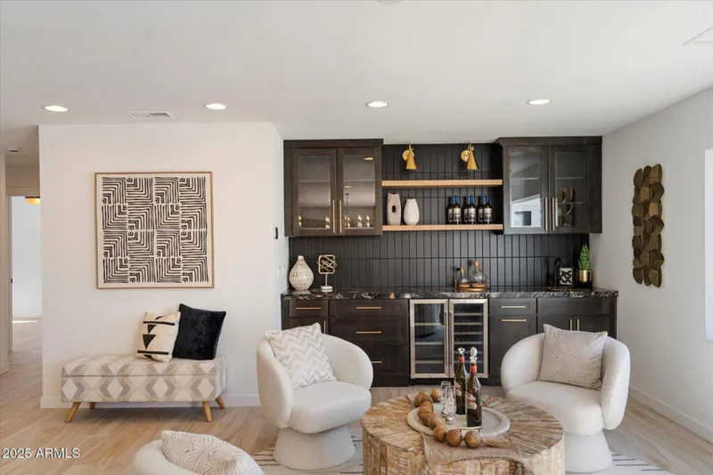 Modern home bar with dark cabinets, wine fridge, and two white accent chairs around a wood stump coffee table