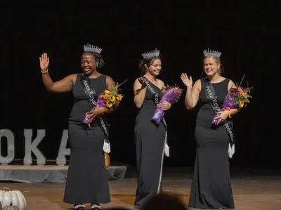 Three women in black gowns with crowns and sashes wave and hold bouquets on a stage