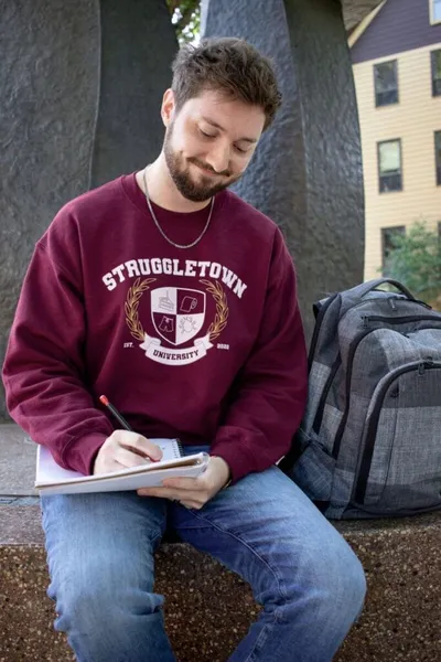 Young bearded man in a maroon sweatshirt writing in a notebook outdoors with a backpack beside him