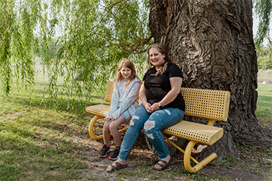 Woman and young girl sitting together on a yellow bench beneath a large tree in a park