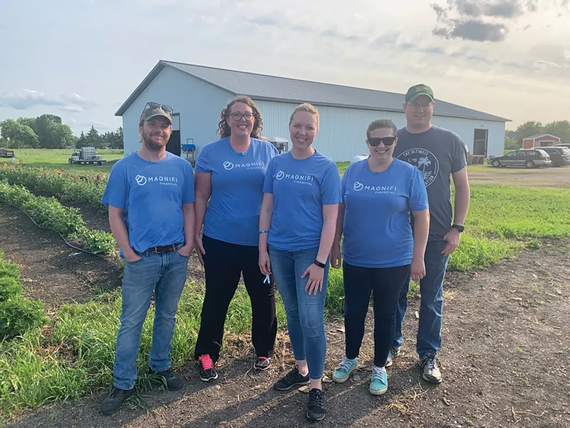 Five people in matching blue Magnifi Financial shirts posing outdoors near a farm building