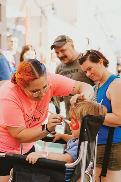 Woman with red hair face-painting a young child at an outdoor community event