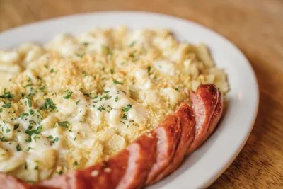 White plate of mac and cheese topped with breadcrumbs and herbs, rimmed with sliced smoked sausage
