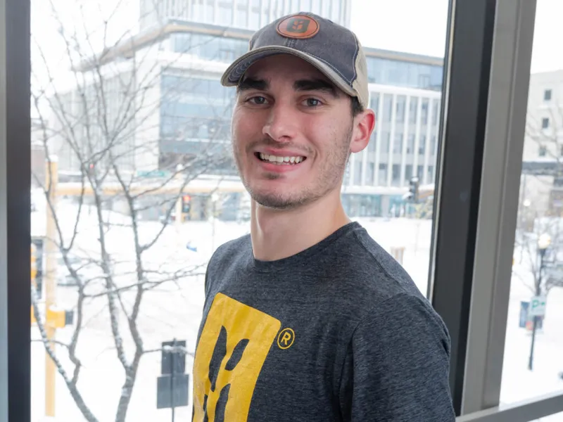 Smiling young man in a gray T-shirt and trucker cap beside a snowy urban window