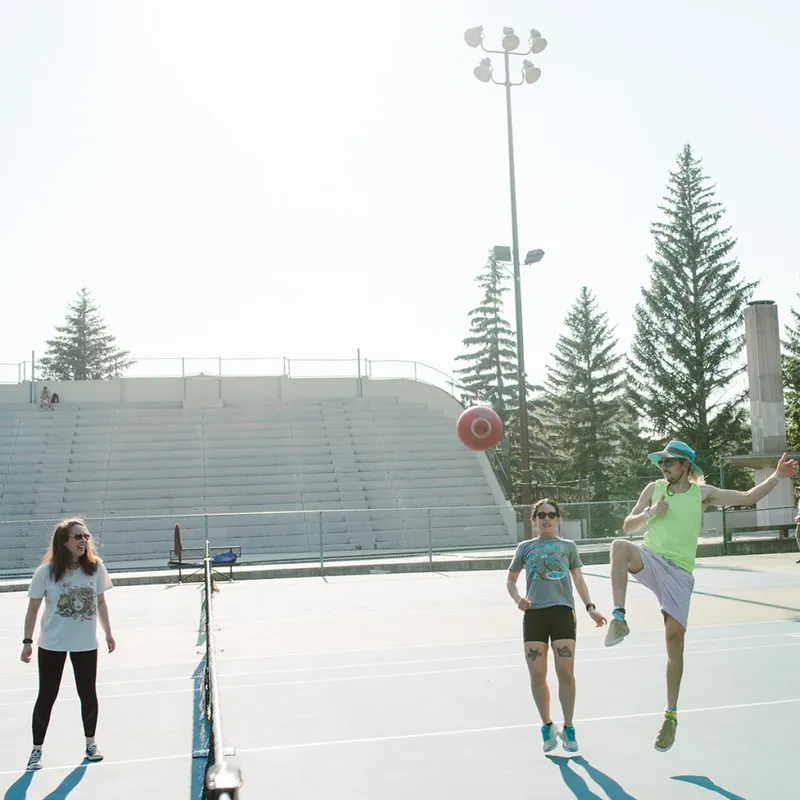 Three people playing a ball sport on an outdoor tennis court, one mid-kick