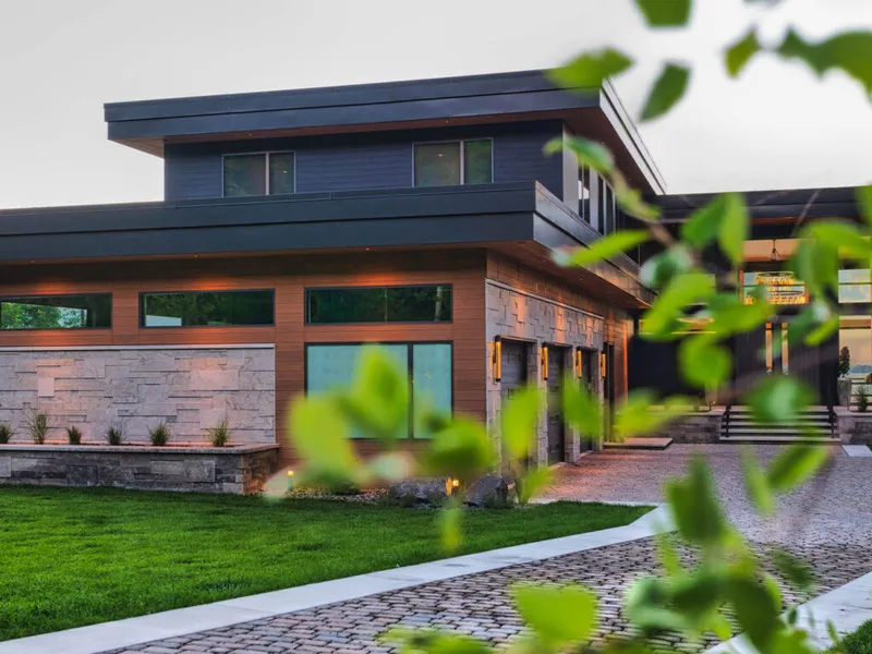 Modern two-story home with stone and wood siding, warm accent lighting, and a cobblestone driveway at dusk