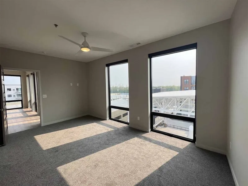 Empty carpeted room with ceiling fan and tall black-framed windows overlooking rooftops