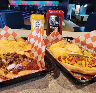 Two loaded hot dogs with chili and toppings beside crinkle-cut chips, ketchup and mustard on a bowling alley table