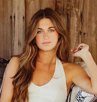 Young woman with long wavy brown hair in a white tank top against a wooden wall