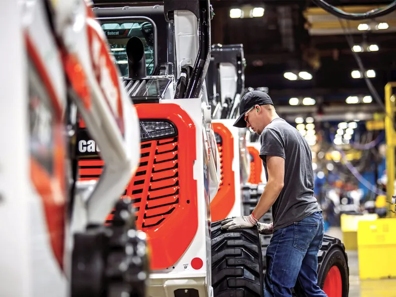 Worker in gloves and cap inspecting a tire on a red-and-white compact loader on a factory floor