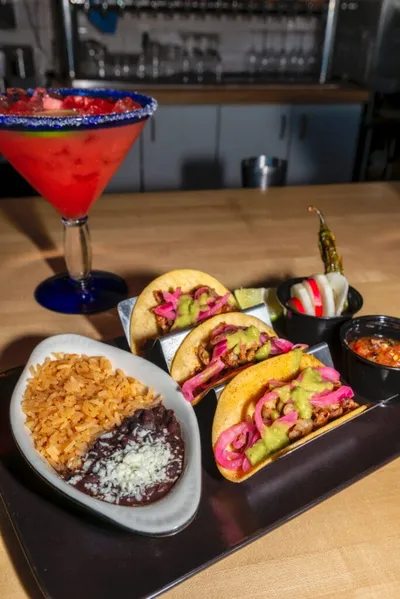 Three street tacos with guacamole and pickled onions, rice and black beans, and a red margarita on a wooden table