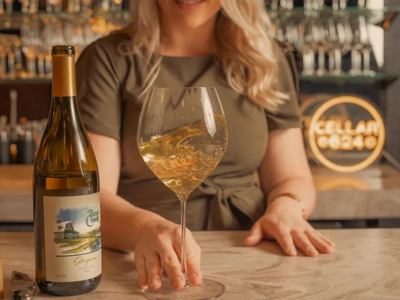 Bartender holds a white wine glass beside a Bear Creek Winery bottle at a bar