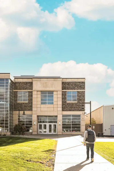 Person with backpack walking toward a two-story brick campus building under a partly cloudy sky