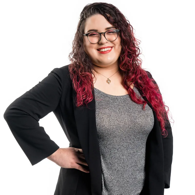 Smiling woman with red-dipped curly hair, glasses, and black blazer, hand on hip