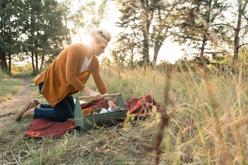 Woman in mustard cardigan kneeling on a red blanket in a grassy meadow, reaching into a green supply caddy at golden hour