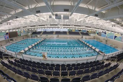 Indoor Olympic-sized swimming pool with lane lines and stadium seating under a large arched roof