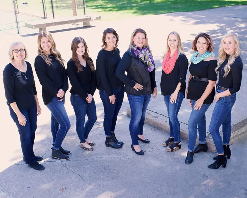 Eight women in black tops and jeans posing together outdoors on a sunny day
