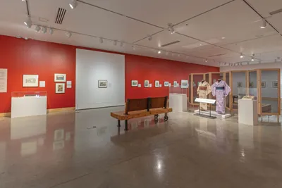 Bright museum gallery with red accent walls, framed artwork, a wooden bench, and two traditional kimono garments on display