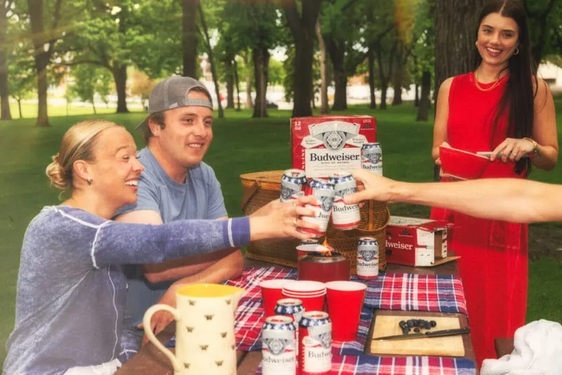 Three people toasting Budweiser cans at a plaid-covered picnic table in a park