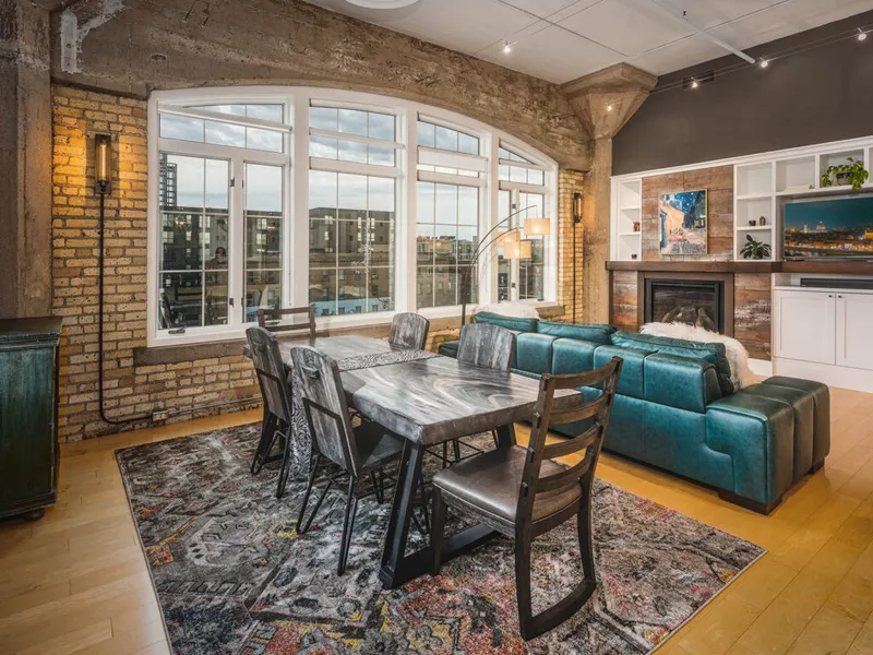 Industrial loft dining area with exposed brick, arched windows, and teal sectional sofa near a fireplace
