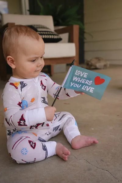 Baby in printed pajamas sitting on a porch holding a teal 'Love Where You Live' flag